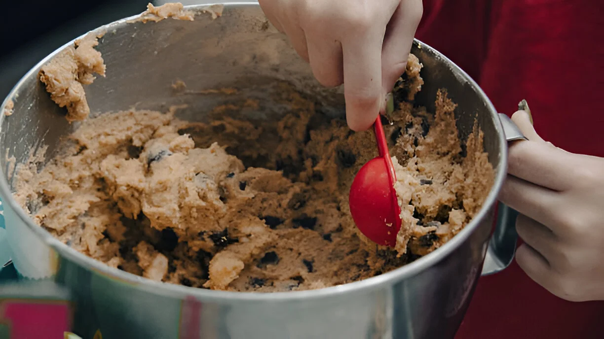 A bowl of homemade protein cookie dough with chocolate chips, nuts, and a sprinkle of sea salt, surrounded by ingredients.